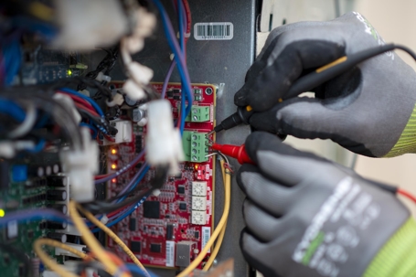 A person with gloves uses a multimeter to test the voltage on a circuit board.